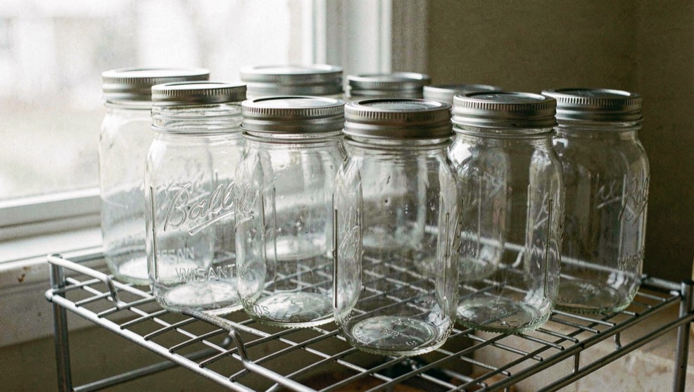 Row of clear glass pint canning jars with two piece metal lids standing on a wire canner rack, empty and dry, in flat daylight from a window