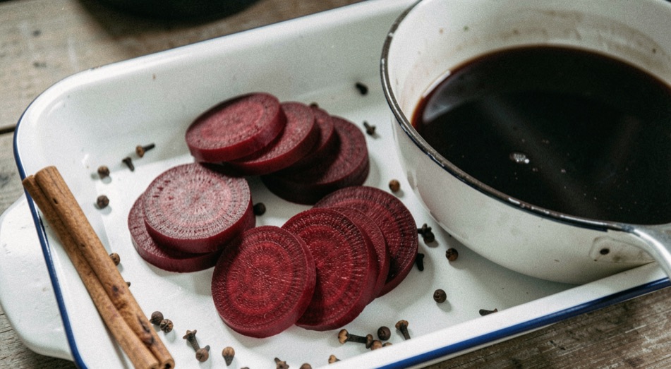 Deep red beets sliced into even rounds on a white enamel tray beside a pan of dark liquid, whole cloves and a cinnamon stick resting on the tray edge