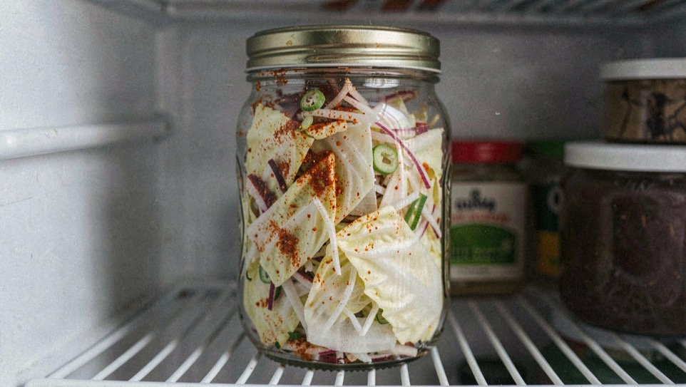 Coarsely cut napa cabbage leaves mixed with red chilli powder, sliced spring onion and shredded radish packed into a clear glass jar standing on a fridge shelf
